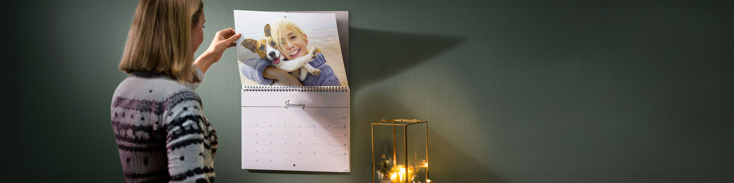 A woman hangs a CEWE photo calendar featuring a joyful photo of her and her dog. A lovely lantern light illuminates the room.